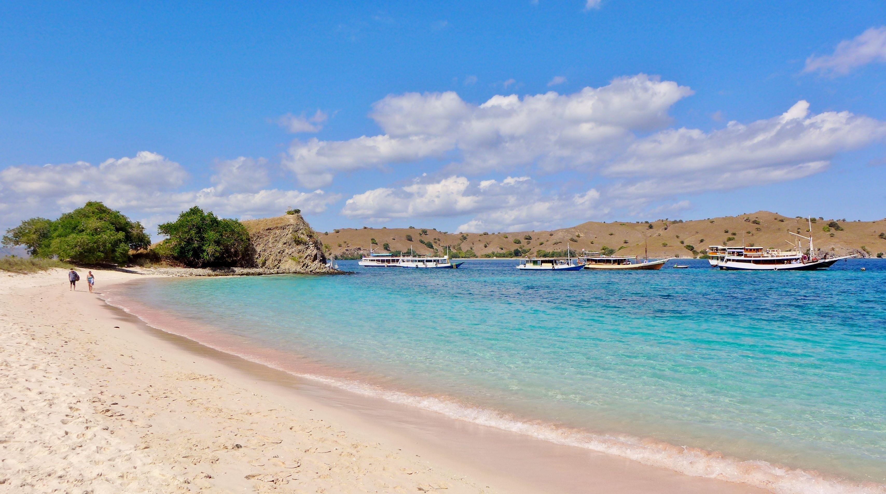 Pink-tinged sand meets turquoise shallows at Pink Beach on Komodo Island, with dive boats moored over the fringing coral garden.